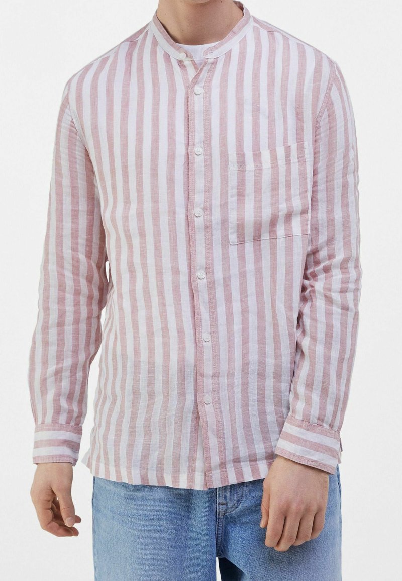 Mand iført lyserød og hvidt vertikalt stribet skjorte med kinesisk krave og blå jeans, stående mod en hvid baggrund.