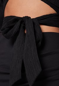 Haut noir avec un tissu texturé et froissé, orné d'un nœud noué à la taille. Jupe noire lisse et ajustée en dessous.