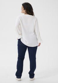 Weiße Bluse mit langen Ärmeln aus strukturiertem Stoff und abgerundetem Saum; kombiniert mit dunkelblauen Jeans und hellen Sneakers.