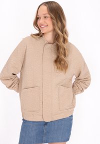Beige steppelt dzseki virágmintával, magas nyakkivágással, hosszú ujjakkal és két elülső zsebbel. Puha tapintású és laza fazonú.