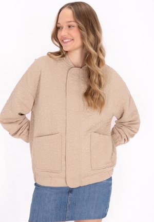 Beige gewatteerde jas met bloemenpatroon, hoge hals, lange mouwen en twee voorzakken. Zachte textuur en een relaxte pasvorm.