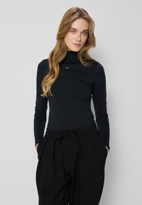 Zwarte langemouwen turtleneck top van een zachte stof, met een subtiele merkaccent op de borst. Gecombineerd met losse zwarte pantalons.