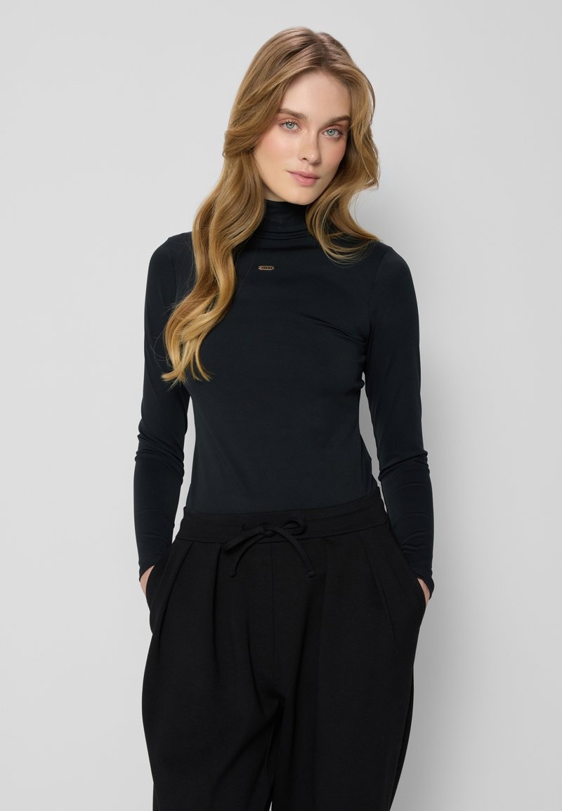 Zwarte langemouwen turtleneck top van een zachte stof, met een subtiele merkaccent op de borst. Gecombineerd met losse zwarte pantalons.