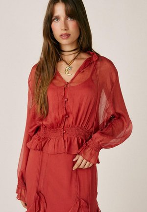 Mujer con blusa roja transparente de manga larga con botones y cintura con volantes, combinada con una falda roja a juego y un collar en forma de luna.