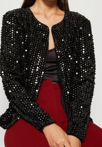 Svart paljetterad jacka med rund halsringning, texturerad sammetfinish och figurnära design. Buren över en svart topp och röda byxor.