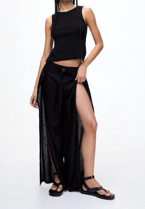 Kvinna som bär en svart ärmlös topp, vida svarta byxor med höga sidoklipp och svarta platta sandaler mot vit bakgrund.