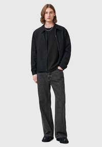 Chaqueta negra ligera con cremallera en la parte delantera, combinada con una camiseta negra y jeans grises holgados. Zapatos negros completan el atuendo.