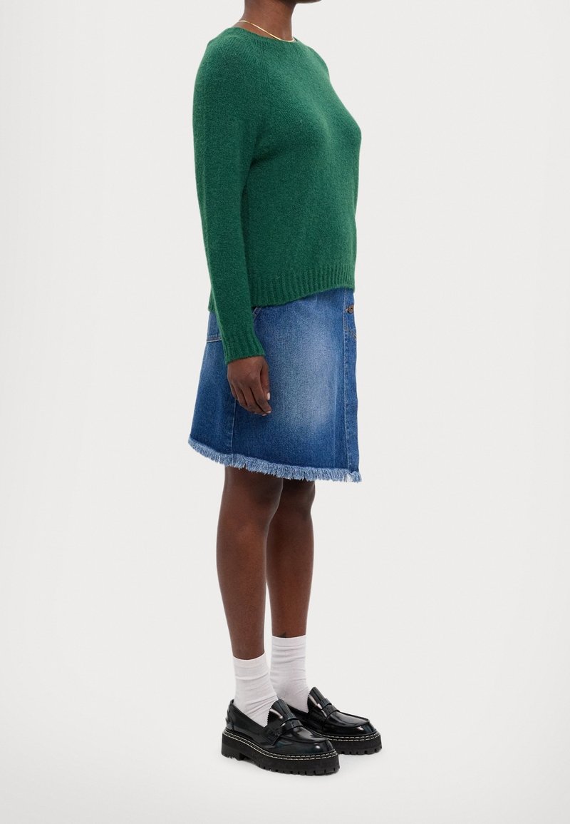 Personne portant un pull en maille vert, une jupe en jean effilochée au genou, des chaussettes blanches et des mocassins noirs épais, debout de profil sur un fond blanc.