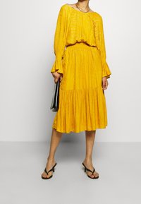 Yellow, textured midi dress with long sleeves and flared cuffs. Features a smocked waistband and a gathered, tiered skirt.
