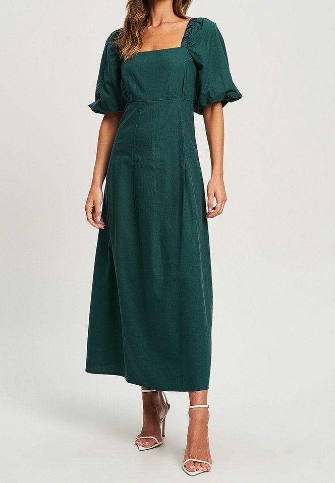 Femme portant une robe vert foncé longue jusqu'aux chevilles avec des manches courtes bouffantes et un décolleté carré, assortie à des sandales blanches à talons hauts.