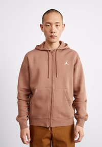 Sudadera marrón con cremallera y capucha ajustable. Material suave, bolsillos tipo canguro y un logo blanco en el lado izquierdo del pecho. Diseño simple y casual.