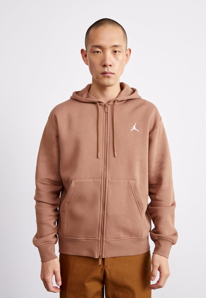 Sudadera marrón con cremallera y capucha ajustable. Material suave, bolsillos tipo canguro y un logo blanco en el lado izquierdo del pecho. Diseño simple y casual.