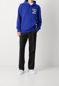 Blauwe hoodie met een kangoeroezak en een capuchon met trekkoord, voorzien van een wit "Calvin Klein Jeans" logo, gecombineerd met zwarte broeken en witte sneakers.