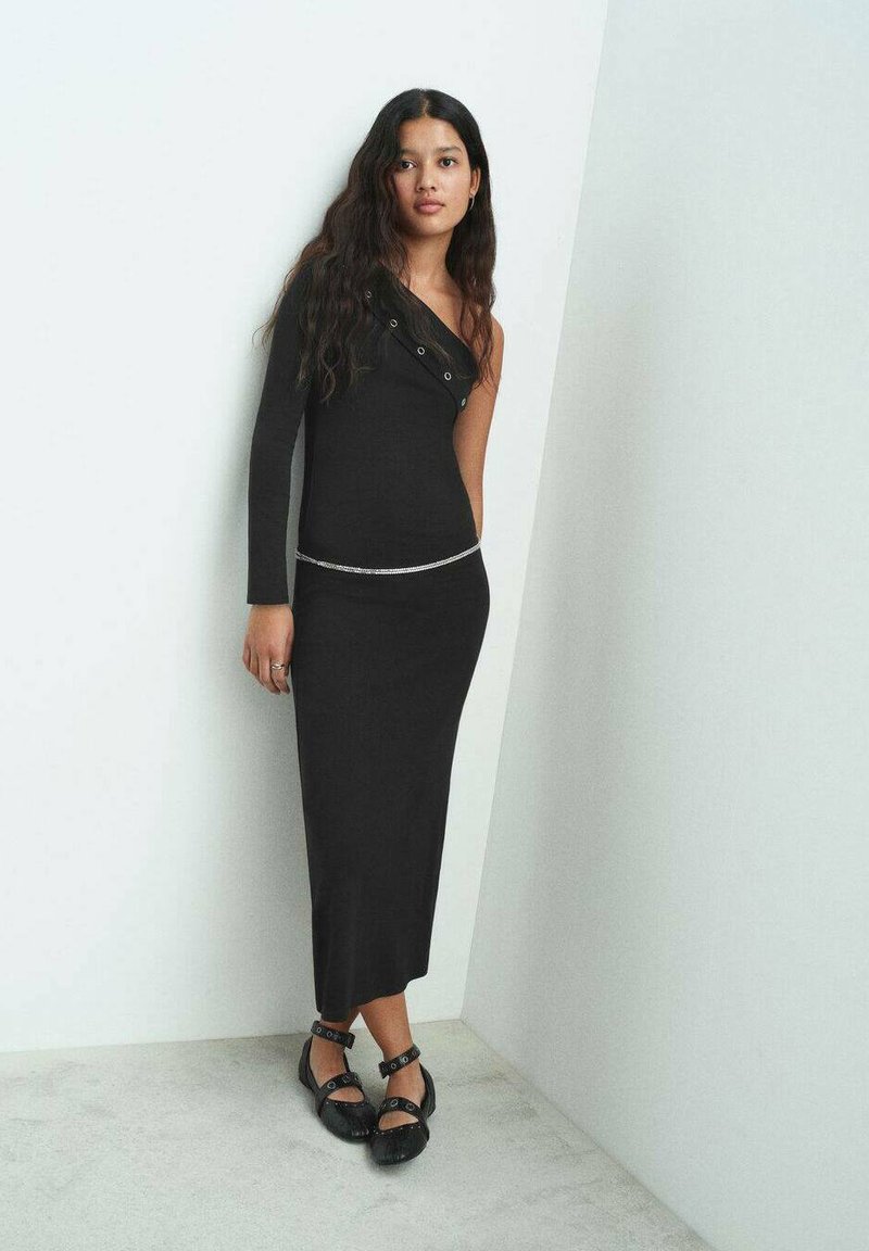 Robe noire ajustée avec un design à une épaule, ornée de détails à boutons-pression et d'une fine ceinture en argent. Porte des chaussures noires à lanières.