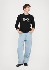 Fekete hosszú ujjú póló fehér "EA7 Emporio Armani" logóval, világoskék, bő szárú farmernadrággal és fekete cipővel párosítva. Egyszerű, hétköznapi öltözet.