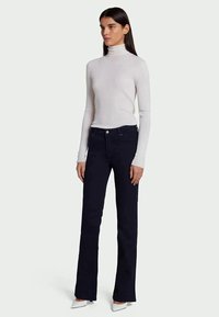 Mulher de pé, de frente para a esquerda, vestindo um suéter branco de gola alta, jeans escuros à boca de sino e sapatos brancos de bico fino.