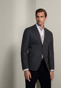 Blazer di lana grigia con revers, chiusura a un bottone e due tasche frontali, indossato sopra una camicia chiara e un maglione senza maniche beige.