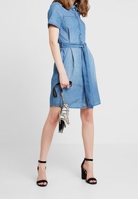 Denim-Kleid in Hellblau mit kurzen Ärmeln, durchgehender Knopfleiste, passendem Gürtel und seitlichem Schlitz. Getragen mit schwarzen High Heels und einer gemusterten Tasche.