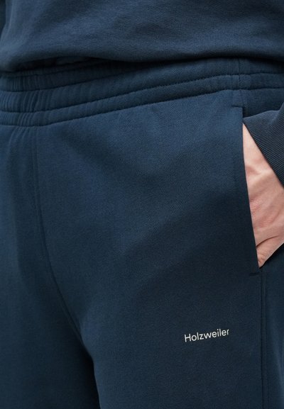 Granatowe spodnie dresowe o miękkiej fakturze, z elastycznym pasem, kieszeniami po bokach oraz białym wyhaftowanym logo „Holzweiler”.