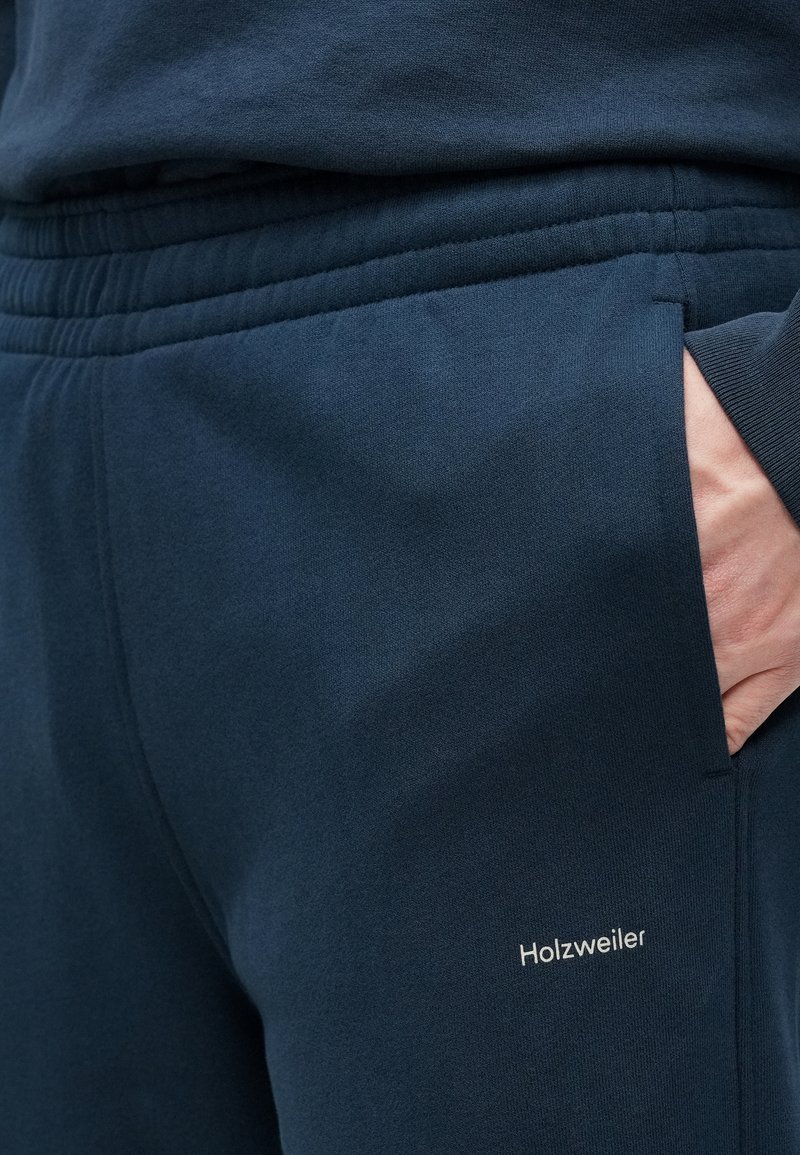 Pantalon de survêtement bleu marine avec une texture douce, une taille élastique, des poches latérales et le logo « Holzweiler » brodé en blanc.