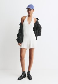 Robe halter blanche en tissu texturé ; veste noire oversize ; casquette bleue ; mocassins noirs associés à des chaussettes foncées. Design simple et moderne.