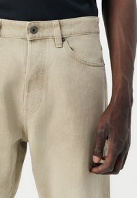 Los pantalones vaqueros de mezclilla beige cuentan con un cierre de botón, bolsillos frontales y costuras visibles. La tela tiene una textura suave y un brillo sutil.