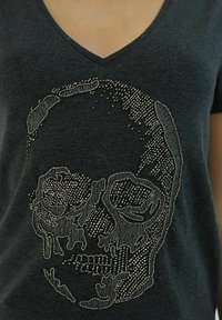Tmavě šedé tričko s výstřihem do V s designem lebky vyrobeným z kovových stříbrných hřebíčků, zdůrazňujícími složité detaily a texturu.