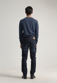 Camisa de manga larga azul oscuro, combinada con jeans de mezclilla oscuros. Los jeans tienen cinco bolsillos y un parche de cuero marrón en la parte posterior. Zapatos negros.