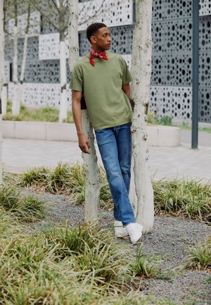 T-shirt verde in cotone, jeans blu, bandana rossa e sneakers bianche. Il modello si appoggia a degli alberi di betulla in un'area esterna paesaggistica.