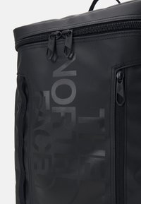 Gros plan d'un coin de sac à dos noir avec double fermeture éclair, une poche verticale zippée et un marquage de texte large et discret sur la surface en tissu.