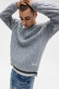 Lys grå cable-knit sweater med ribstrikkede manchetter og kant, lagdelt over en stribet skjorte. Mørkeblå slidte jeans, synlig mærke.