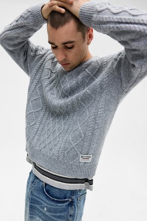 Pull en maille cable gris clair avec des poignets et un ourlet côtelés, superposé à une chemise rayée. Jeans usés bleu marine, marque visible.