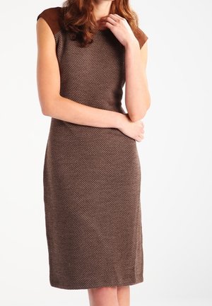 Mujer con vestido marrón texturizado sin mangas hasta la rodilla, con paneles en los hombros de un marrón más oscuro, de pie con los brazos cruzados y la mano cerca de la barbilla.