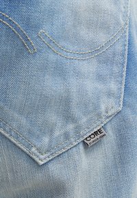 Vaaleansininen denim, jossa on näkyvät ompeleet, kaarevat taskusilmukat ja vyötärönauhassa bränditag, jossa lukee "CORE".