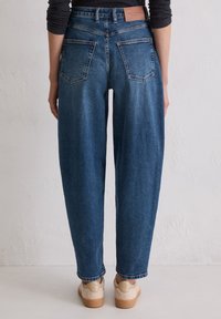 Personne portant un jean en denim bleu taille haute avec des poches arrière, un haut noir et des baskets beiges, se tenant contre un mur texturé clair.