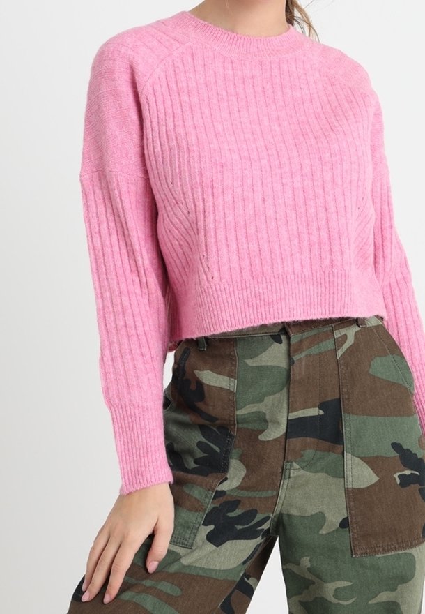 Femme portant un pull court rose côtelé et un pantalon cargo camouflage vert-brun, la main posée sur sa cuisse.