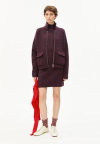 Lila strukturiertes Sweater- und Rock-Set mit hohem Kragen und Reißverschluss; das Model hält ein rotes geripptes Kleidungsstück; weiße Schuhe mit Schnürsenkeln.