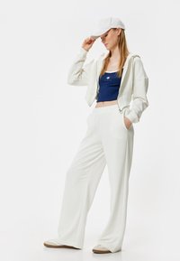 Witte hoodie, marineblauwe crop top met de tekst "PARIS", witte wijde broek en witte pet. Gladde stof en relaxte pasvorm.