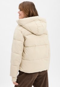 Lichtbeige pufferjack met capuchon, met gestikte panels en een gestructureerd oppervlak. Inclusief een logo patch op de mouw.