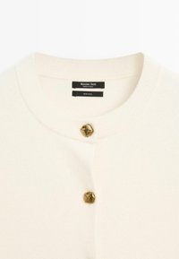 Wollcardigan in Off-White mit rundem Ausschnitt, versehen mit einem markanten, strukturierten goldfarbenen Knopf am Kragen. Zwei sichtbare Markenetiketten.