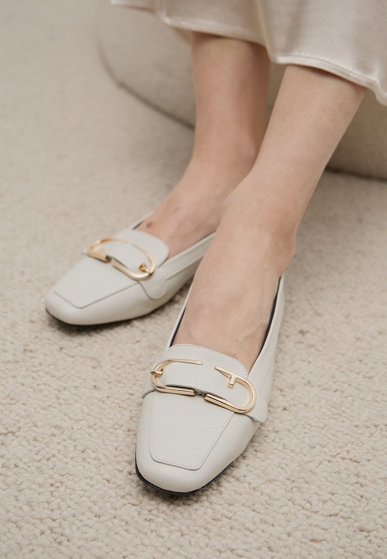 Hellegraue Lederloafer mit quadratischer Zehenform, verziert mit einer goldenen Metallschnalle auf dem Rist. Glatte Textur, minimalistische Designelemente.