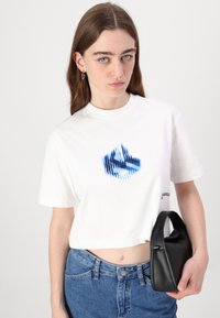 Bela skrajšana majica s kratkimi rokavi in modrim grafičnim dizajnom, okrogla izrezana v vrat. V kombinaciji z visokim pasom denim kavbojkami in črno torbico.