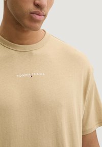 Béžové bavlněné tričko s kulatým výstřihem a vyšívaným logem "TOMMY JEANS" s malým barevným pruhovým detailem na prsou.