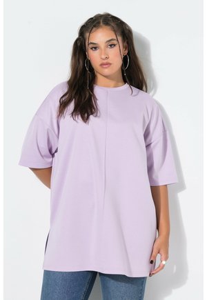 Ung kvinde med langt hår og hoopsøreringe iført en løs lavendel t-shirt og blå jeans, stående mod en ensfarvet hvid baggrund.