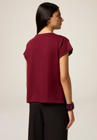 T-shirt bordeaux à manches courtes en coupe décontractée, fabriqué à partir d'un tissu doux. Il présente des manches roulées et un ourlet droit, porté avec un pantalon noir.