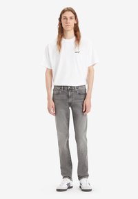 Grå jeans med slim fit, hvit T-skjorte med en liten svart logo, og hvite sneakers med svarte striper. Enkel, uformell stil.