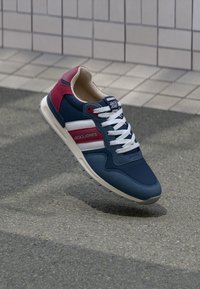 Sneaker Jack & Jones blu navy e rosso con lacci bianchi che galleggia sopra l'asfalto vicino a un muro piastrellato.
