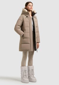 Donna che indossa un cappotto invernale imbottito beige con cappuccio, leggings beige e stivali da neve bianchi isolati, in piedi su uno sfondo neutro.