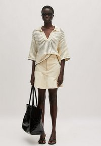 Crème gebreide oversized trui met korte mouwen, gecombineerd met een lichtbeige spijkerrok en een zwarte schoudertas. Zwarte zonnebril maakt de outfit compleet.