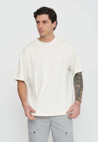 T-shirt overdimensionato di cotone color crema con maniche corte, dotato di un piccolo emblema rotondo sul lato sinistro del petto e una trama liscia.